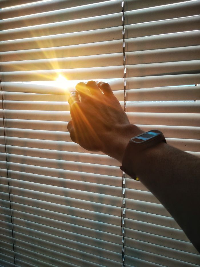 A hand adjusting window blinds, allowing sun rays to pass through in an indoor setting.